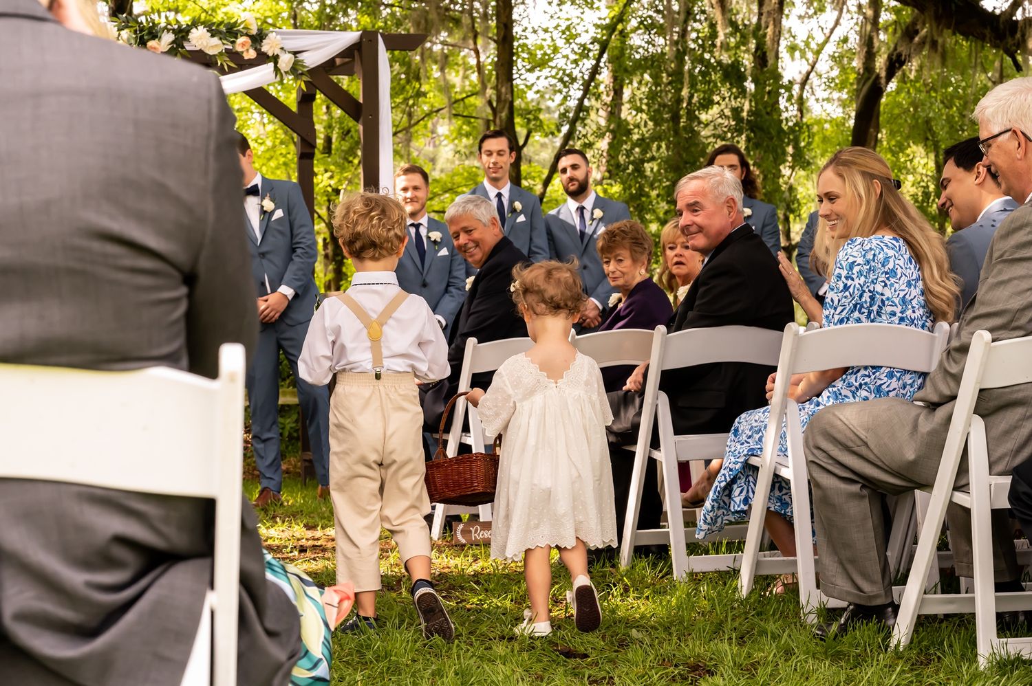 Young flower girl and ring bearer walk down outdoor wedding aisle as guests smile from white folding chairs at garden ceremony.
