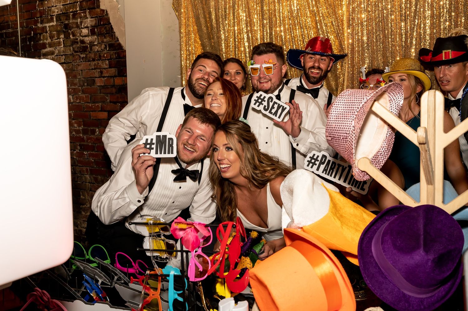 Group of party guests pose with colorful props and accessories in a photo booth setting against a wood-paneled wall.