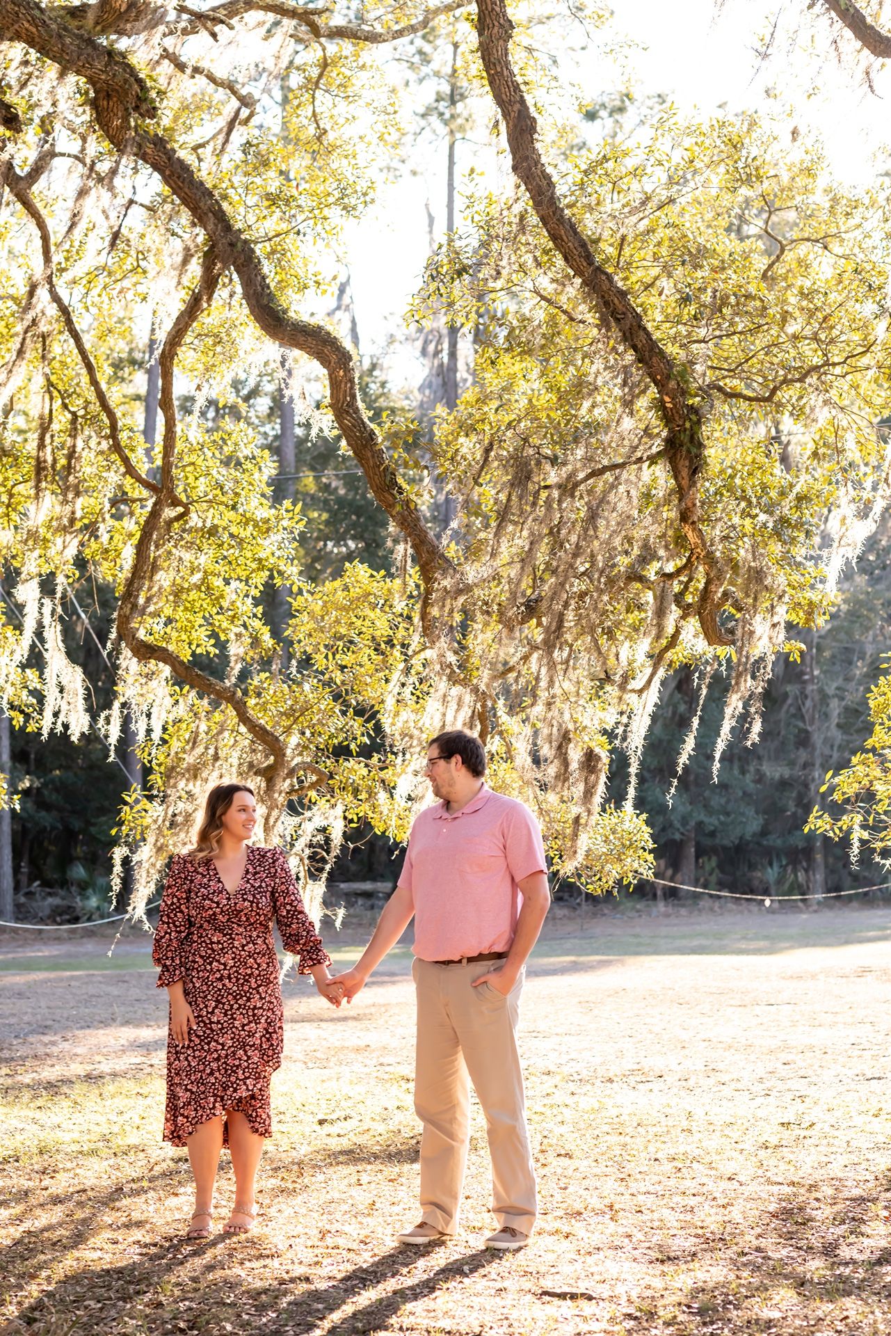 Two people hold hands while standing on fallen leaves under golden autumn trees in a sunlit park.