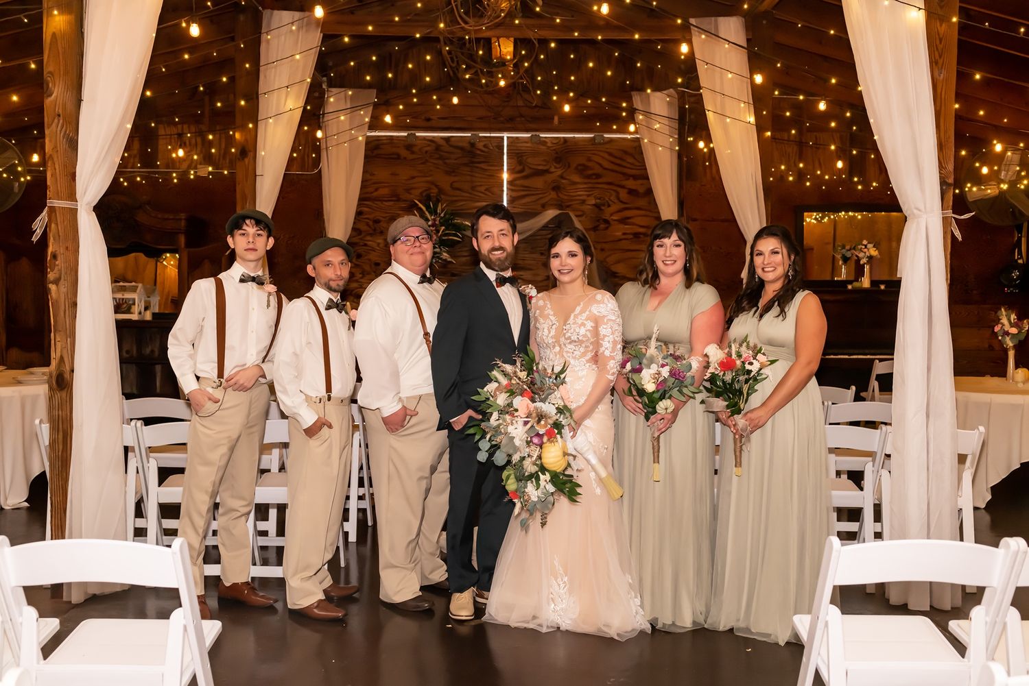 Wedding party and guests stand in an elegant reception hall decorated with white fabric draping and twinkling lights.