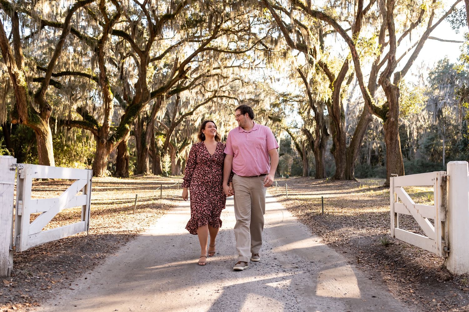 Couple walking hand in hand down a tree-lined dirt path through an oak grove with Spanish moss overhead.