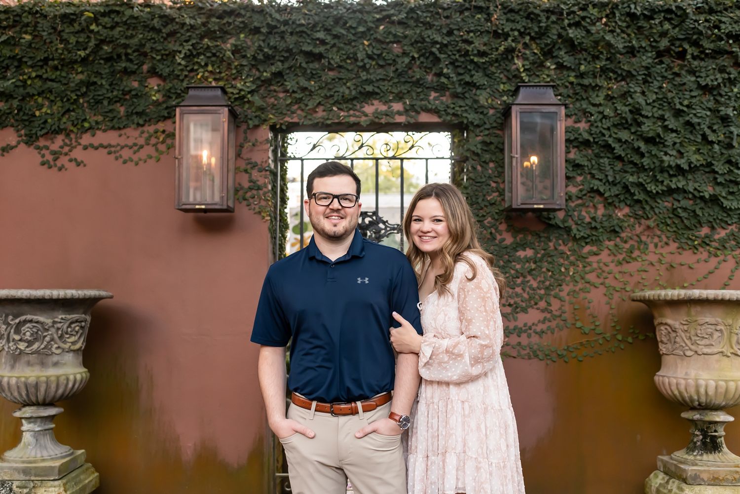 A couple stands in front of a garden wall with ivy, framed by symmetrical lanterns and decorative urns.