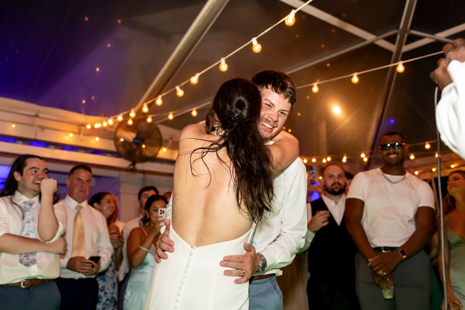 Newlyweds sharing a romantic dance under warm string lights at their wedding reception as guests watch and celebrate.