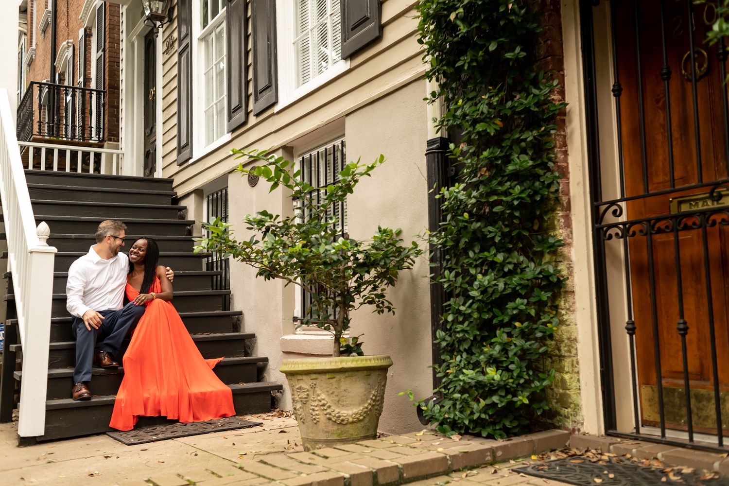 Two people sit on historic brownstone steps in Charleston with orange decor and climbing ivy on the facade.