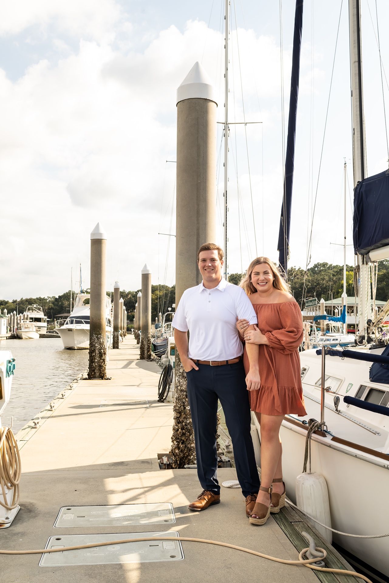 A couple poses together on a marina dock lined with tall white pillars and boats on a sunny day.