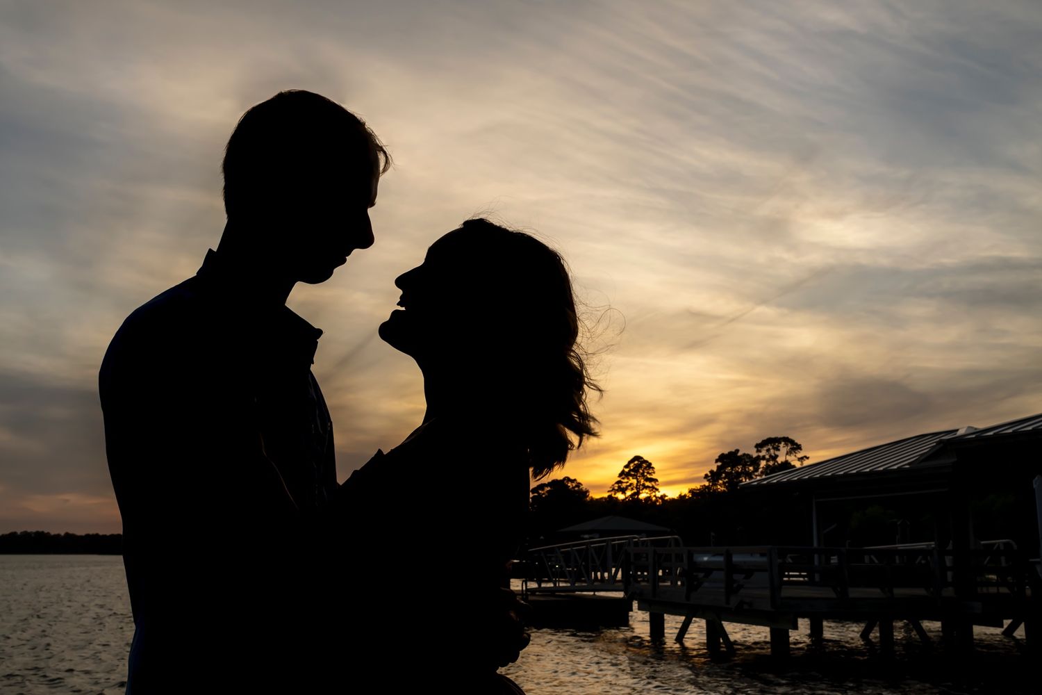 Romantic silhouette of couple embracing by the water during golden sunset with dock in background.