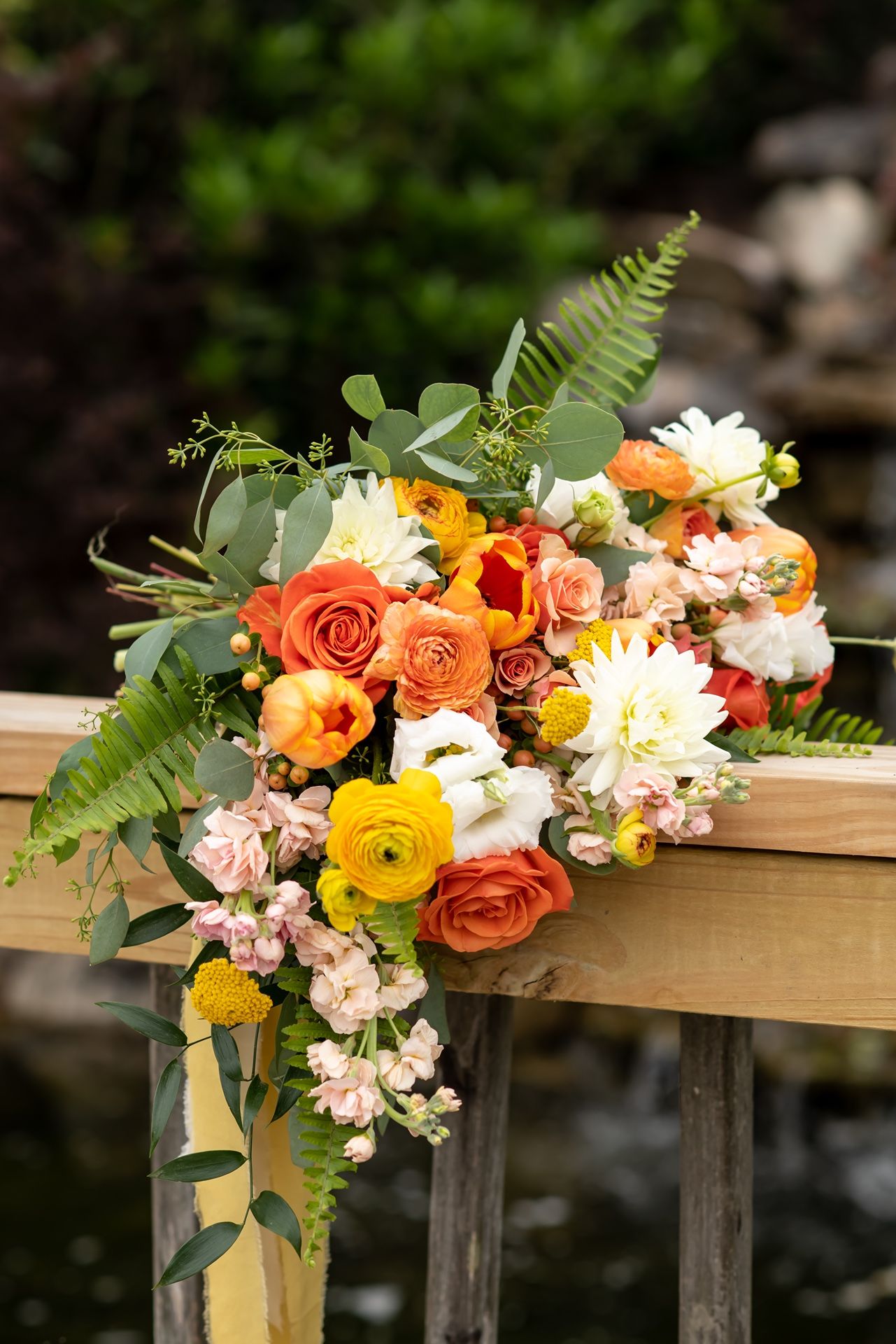 Colorful floral arrangement with orange roses, white blooms and ferns adorns a wooden ledge outdoors in natural lighting.