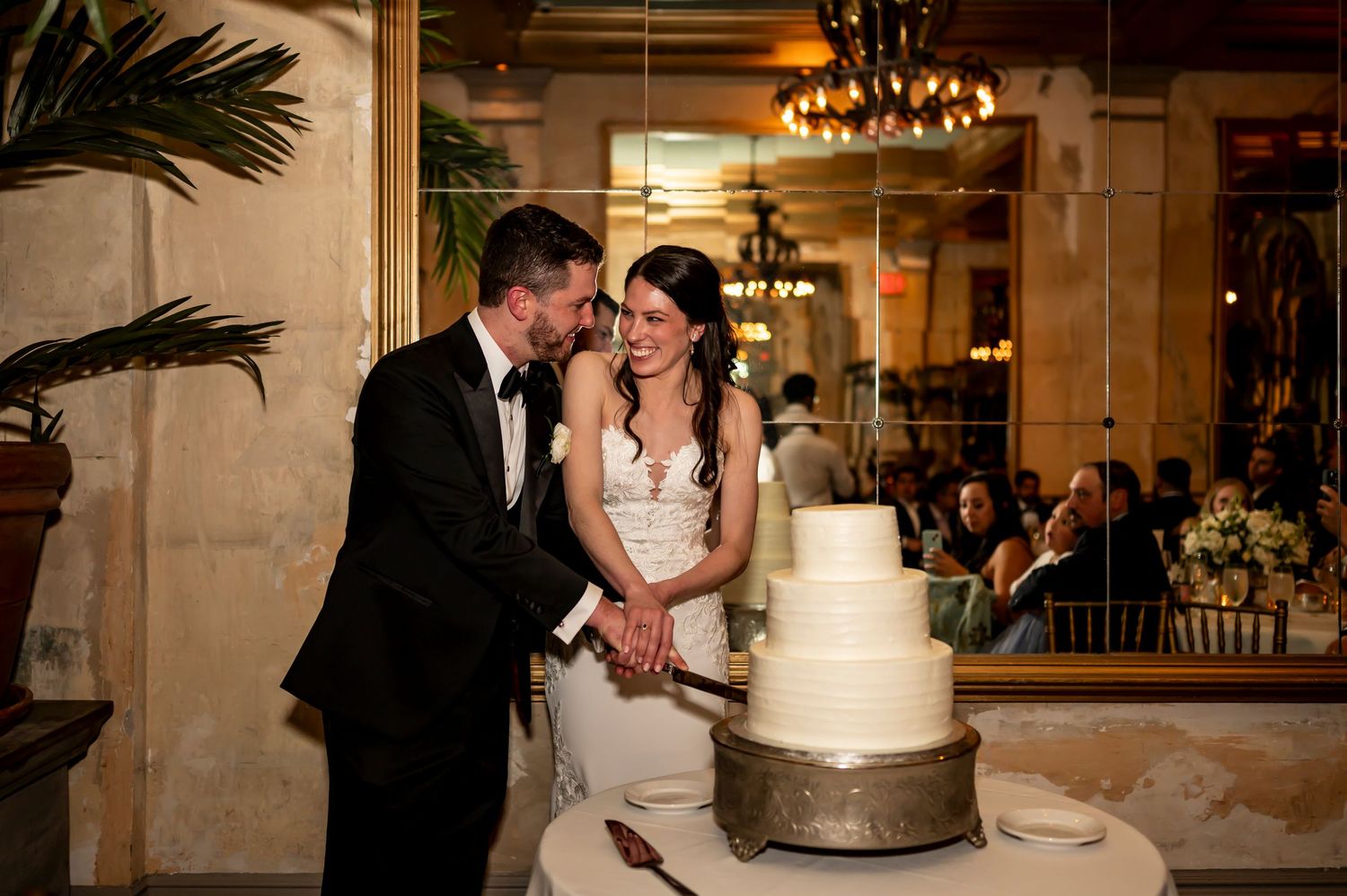 A bride and groom share a tender moment while cutting their three-tiered white wedding cake at an elegant indoor reception.