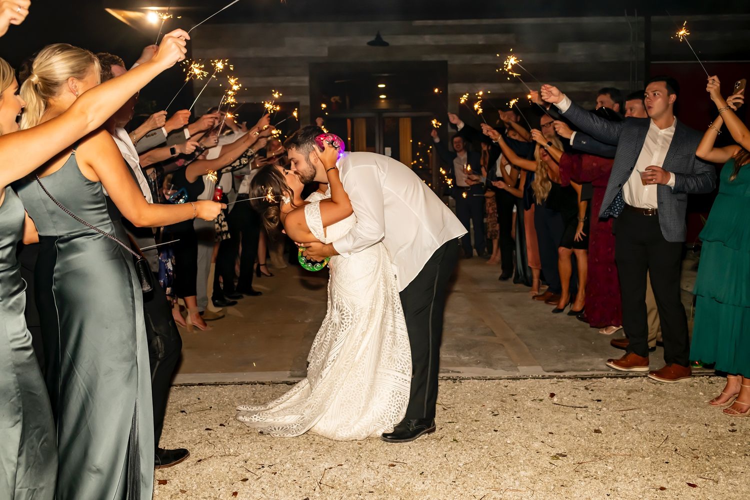 Wedding couple shares romantic kiss during sparkler exit sendoff as guests line pathway holding sparklers at nighttime reception.