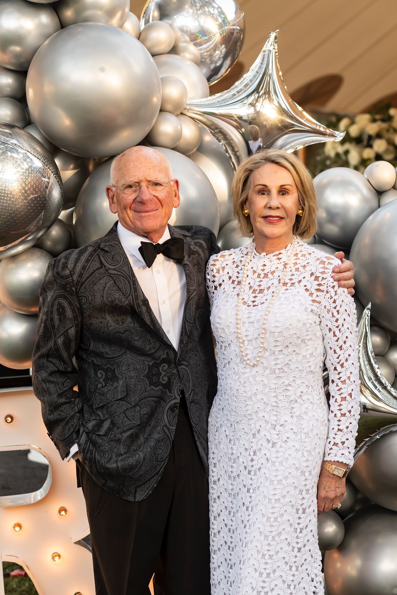 Elegant couple poses in formalwear by silver balloon arch backdrop at festive celebration.
