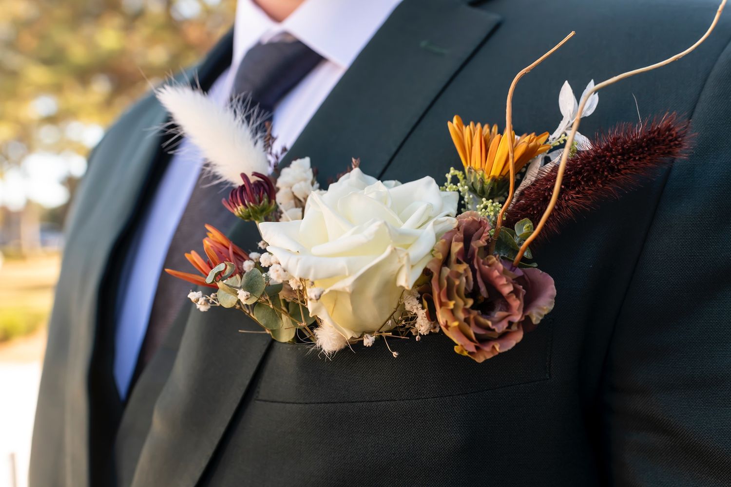 Elegant autumn boutonniere with white rose, orange flowers and feathers pinned to a groom's dark suit lapel.