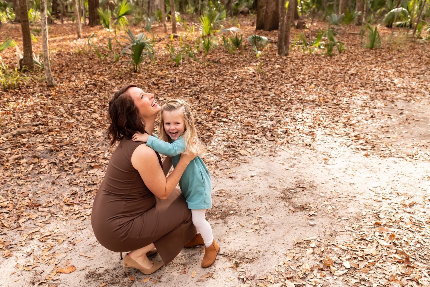 Two people embrace while crouching on a leaf-covered ground in a wooded outdoor setting with filtered sunlight.