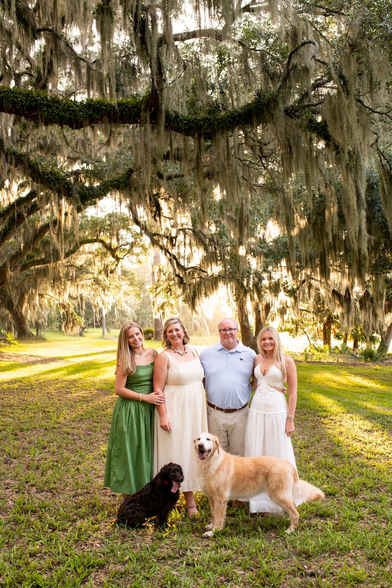 Family of four poses with two dogs under sprawling Spanish moss-draped oak trees in a sunlit Southern landscape.