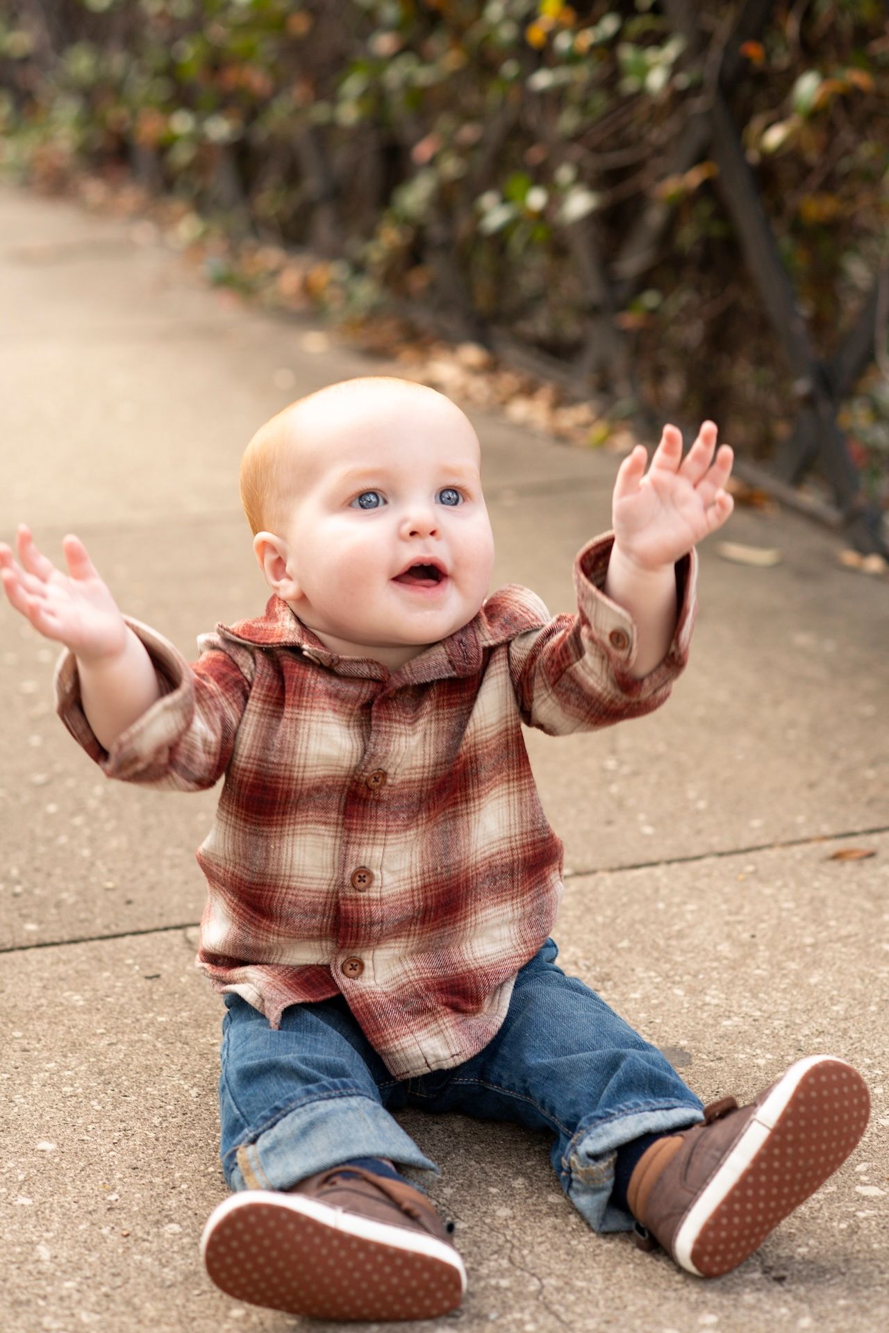 A cheerful baby in a plaid shirt and jeans sits on a path, raising both hands with a joyful expression.