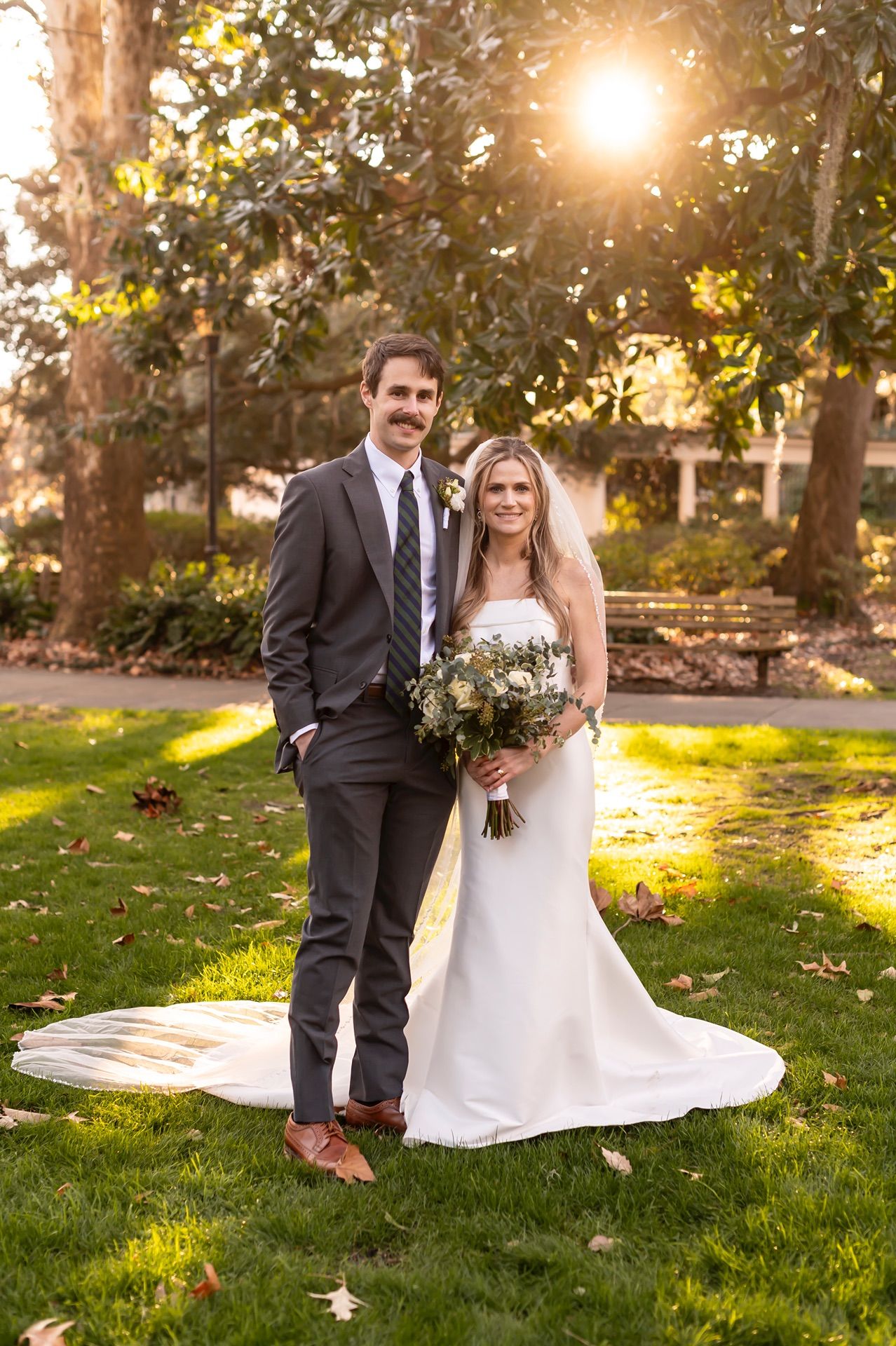 Newlywed couple poses together on green lawn at sunset with beautiful golden sunlight filtering through trees.