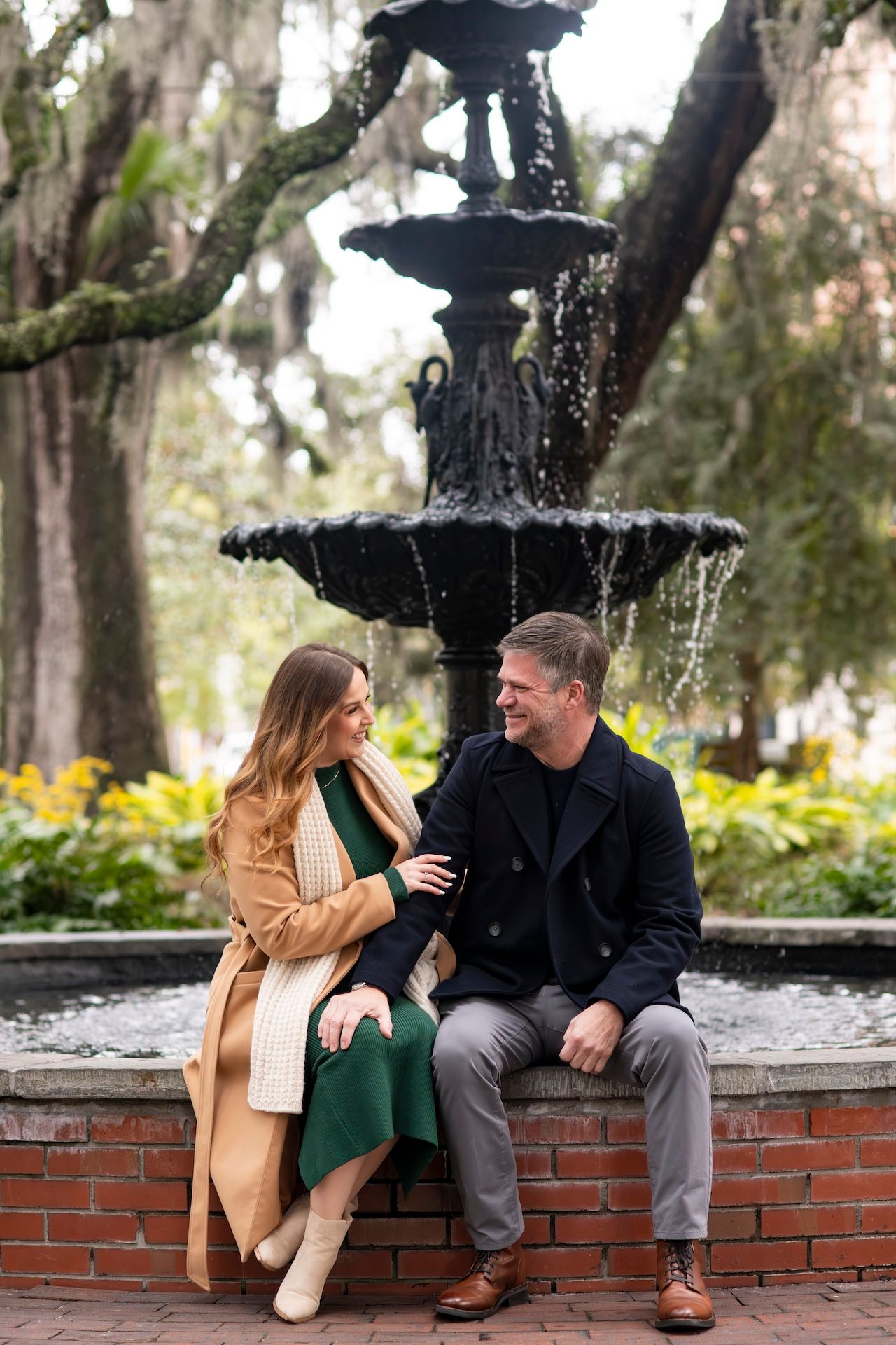 Couple sitting together on brick ledge by ornate black fountain in garden setting with Spanish moss draped trees.