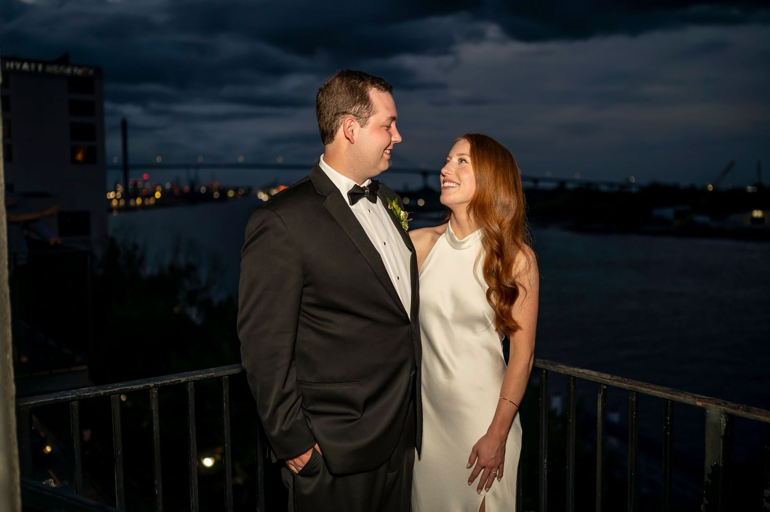 A couple in formal attire stands on a balcony at night with a dark cityscape and water in the background.