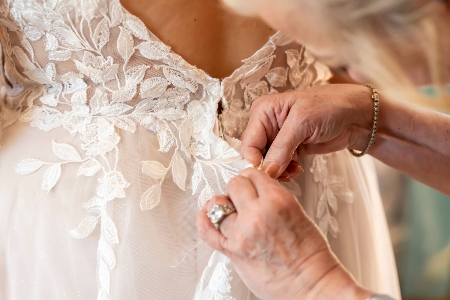 Wedding dress details show delicate lace appliques and buttons being carefully fastened along the back.
