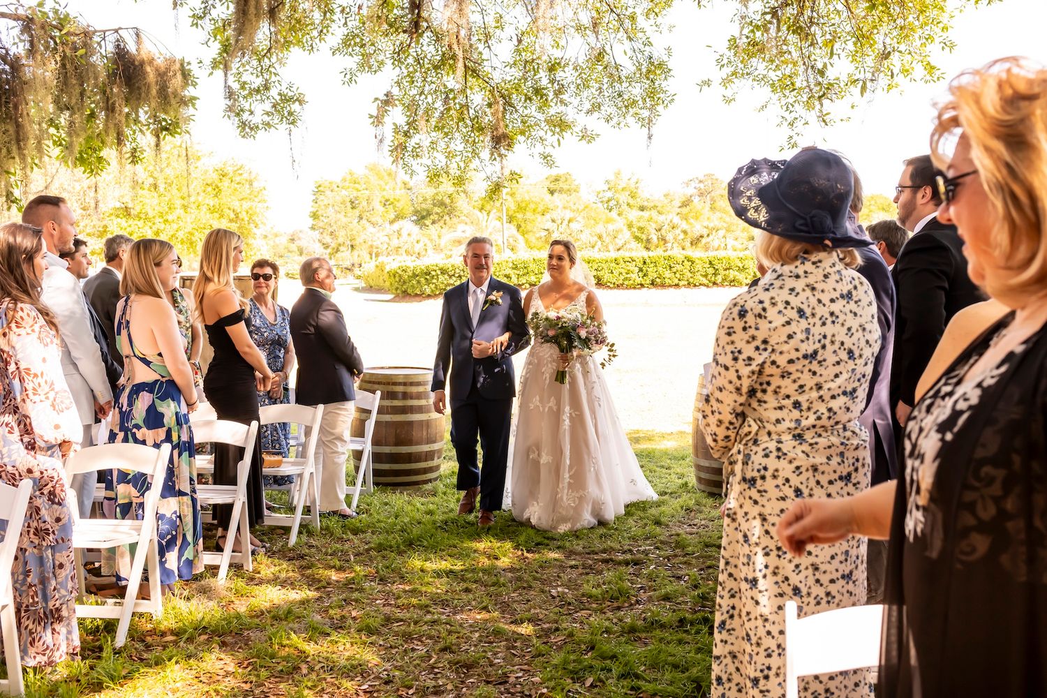 An outdoor wedding ceremony takes place on a sunny day with guests seated on white chairs beside a tranquil waterfront.