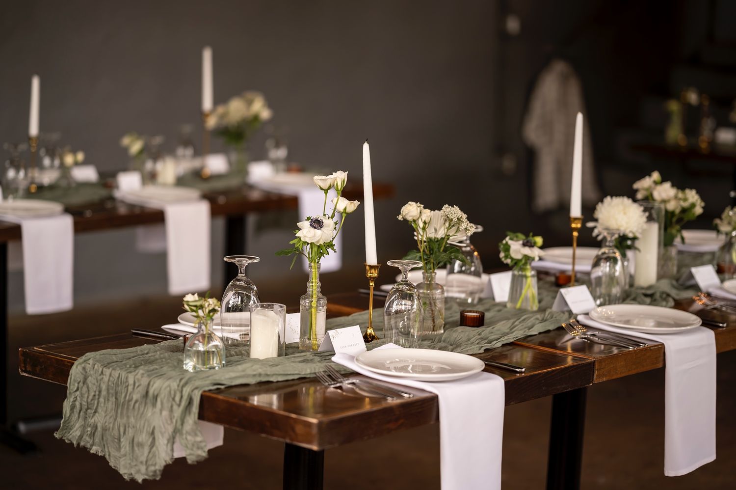 Rustic wedding table setting with white candles, floral centerpieces, and sage green table runners on wooden tables.