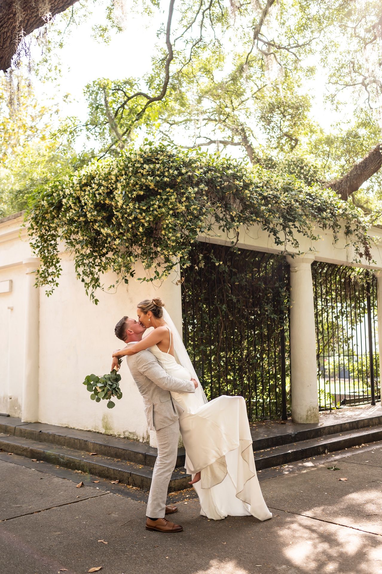 A romantic wedding couple shares kisses under a shady tree-covered archway in a series of intimate outdoor photos.