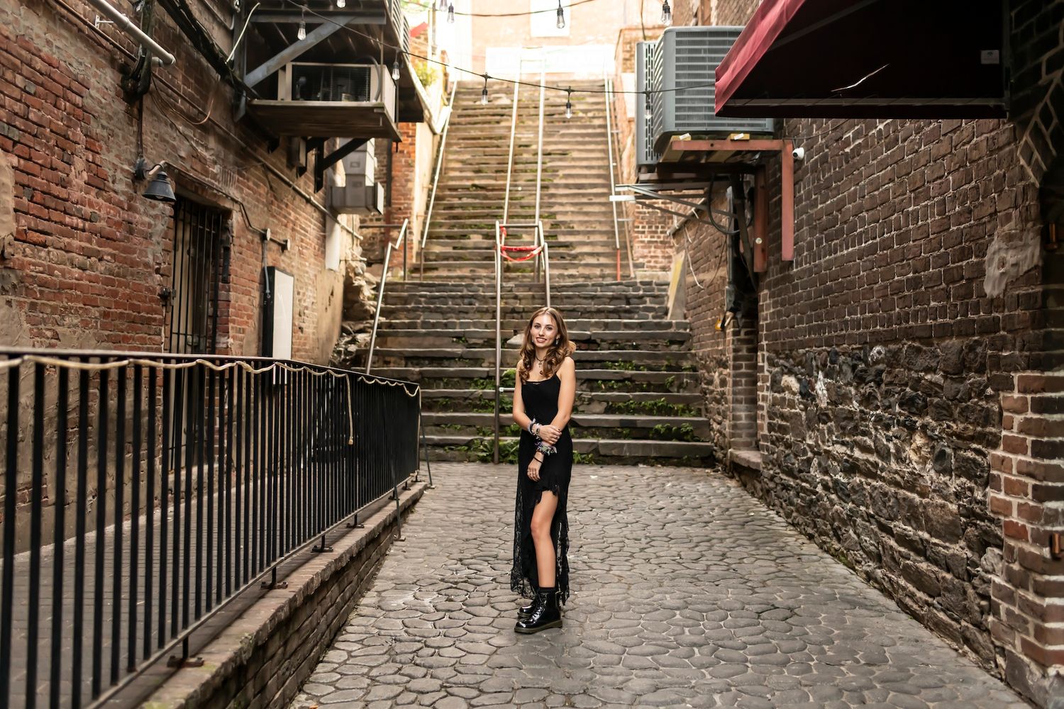A figure in black poses on a cobblestone path framed by brick walls and a large staircase behind them.