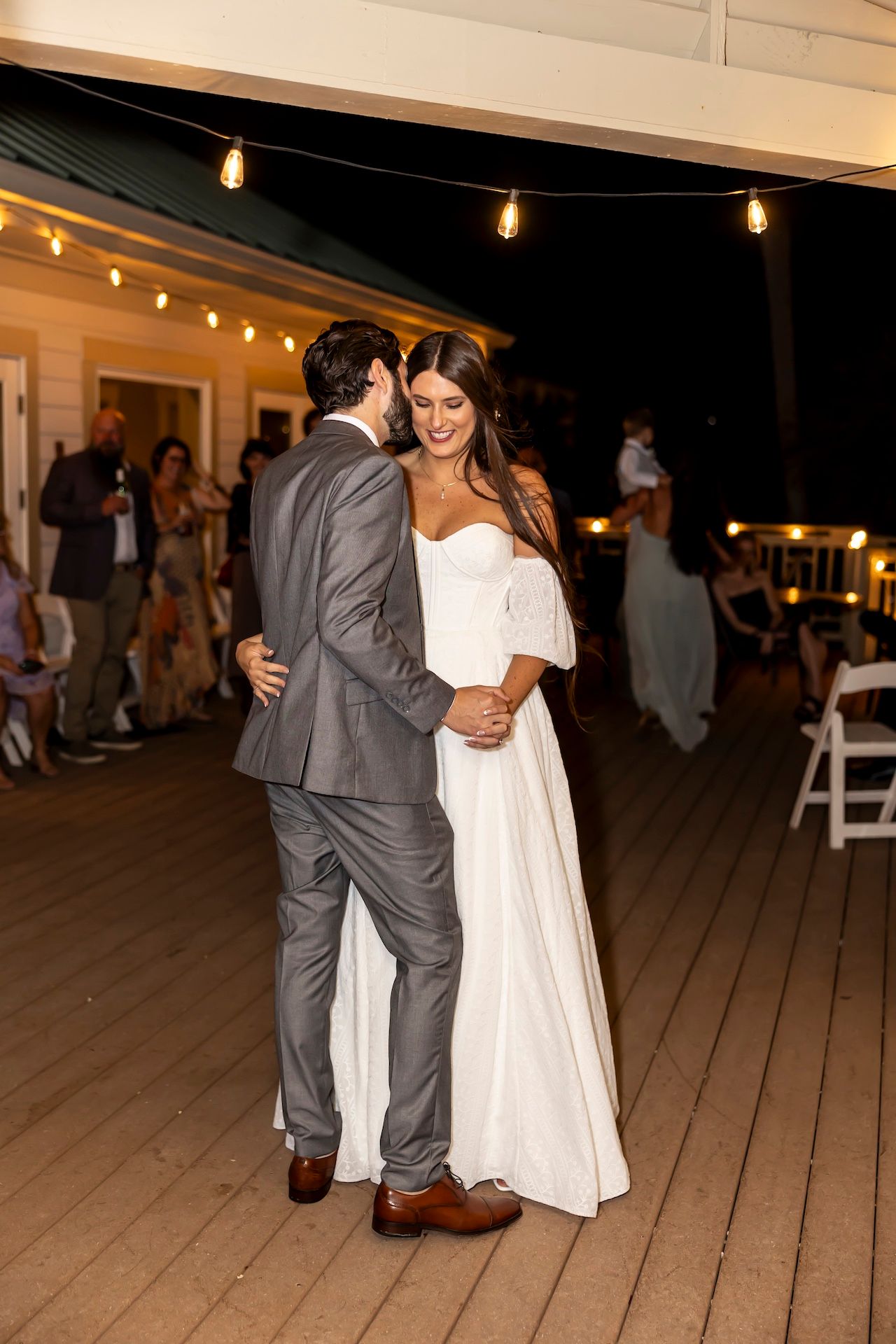 Couple dancing on a wooden deck with string lights glowing above at night during their wedding reception.