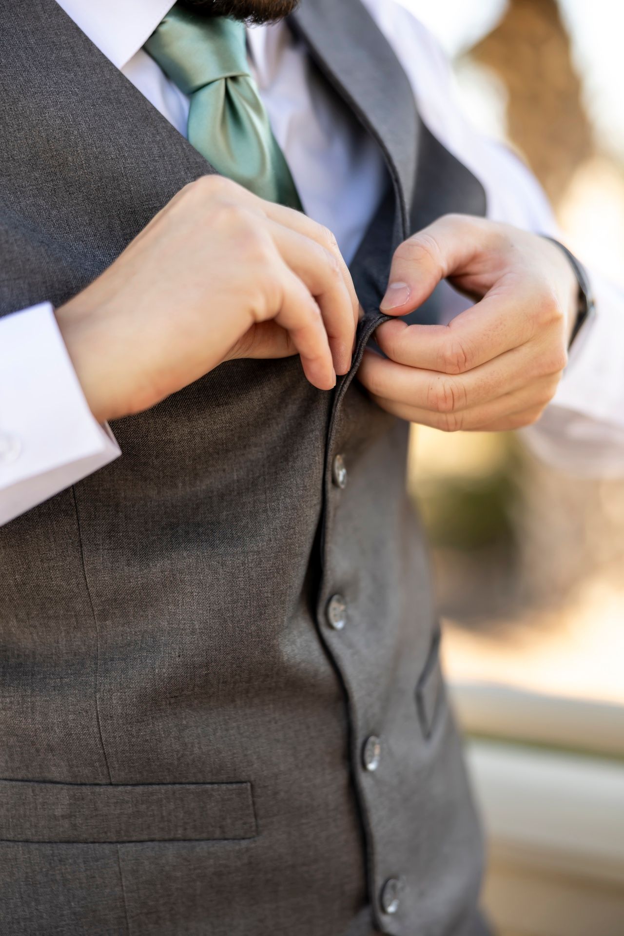 Close-up view of hands buttoning a gray formal vest with mint green tie visible.