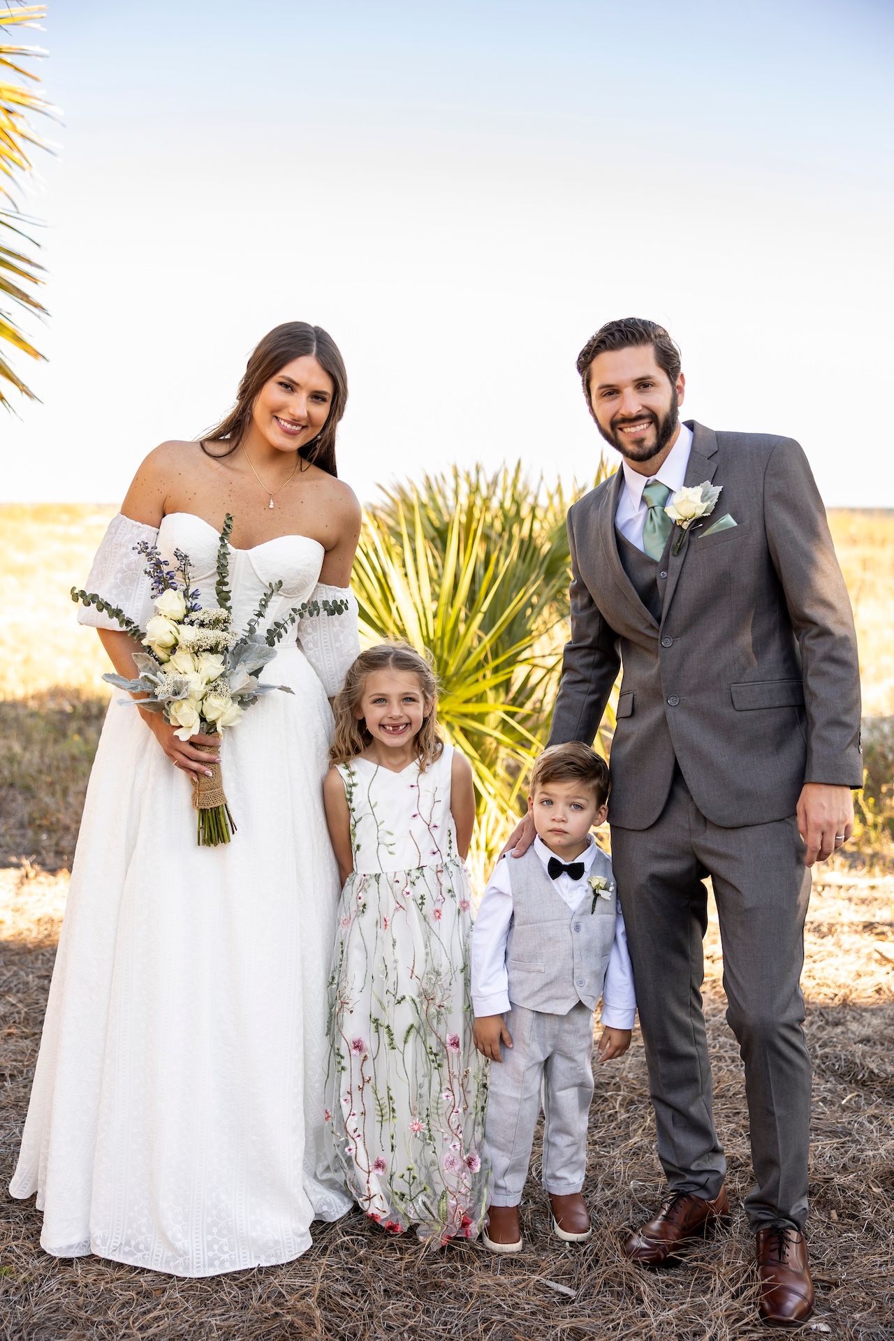 A family in wedding attire poses outdoors with palm trees and desert landscape in the background.