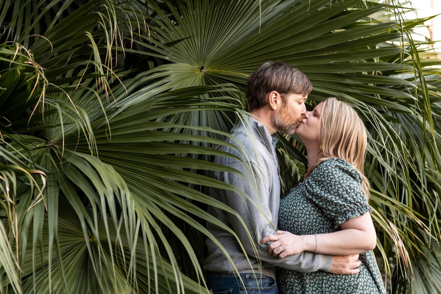 Romantic couple embraces in front of tropical palm fronds on sunny afternoon.