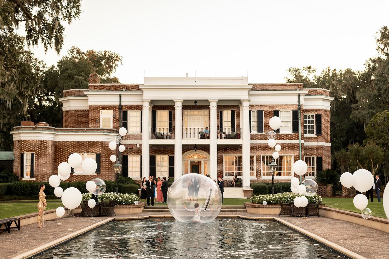 Elegant southern mansion with brick exterior and white columns features a reflecting pool and decorative white balloon decor.