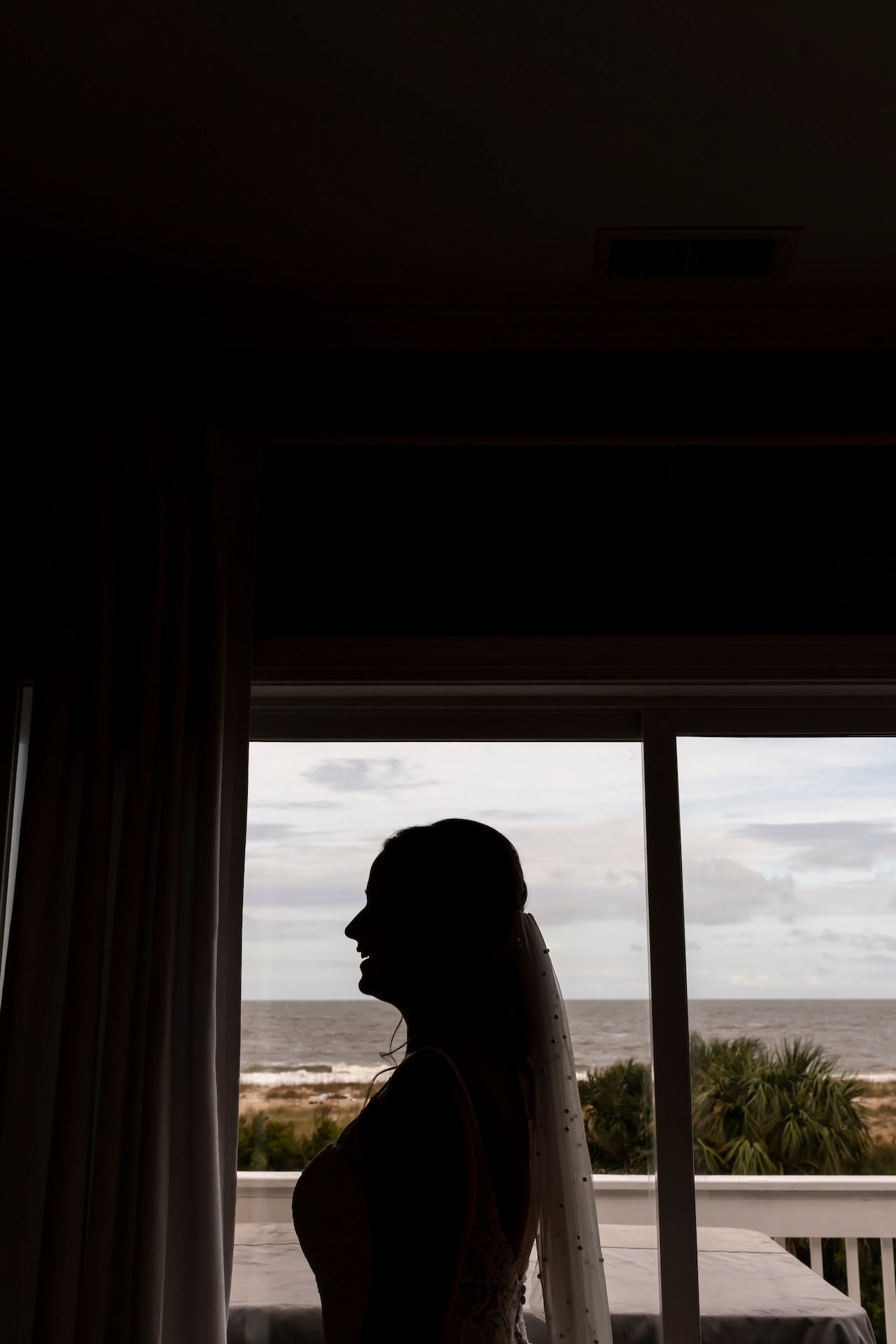 Silhouette by window overlooking beach and ocean, with long veil flowing in seaside resort setting.