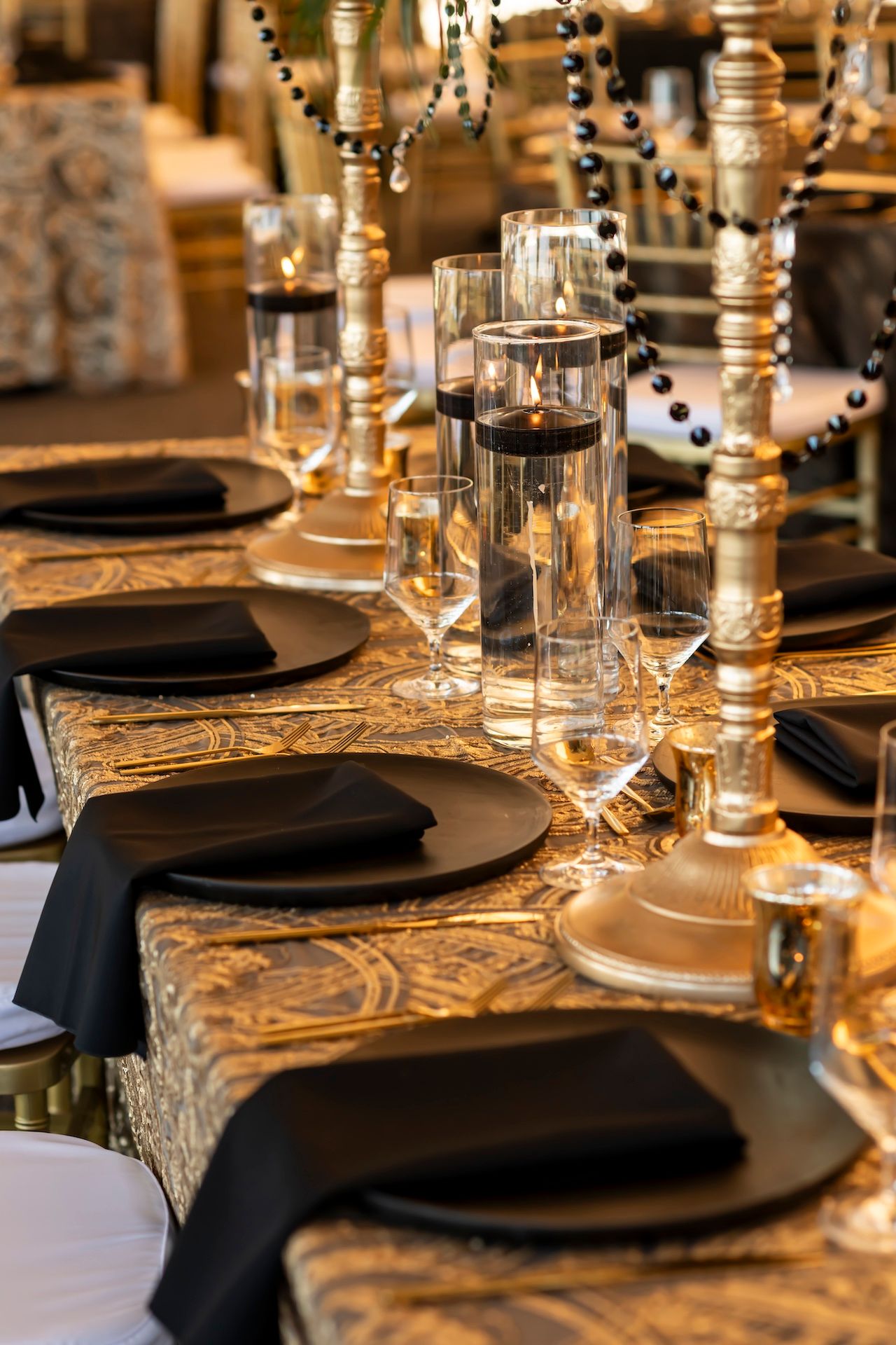 Luxurious table setting with gold tablecloth, black napkins, and crystal candle holders in dramatic lighting.