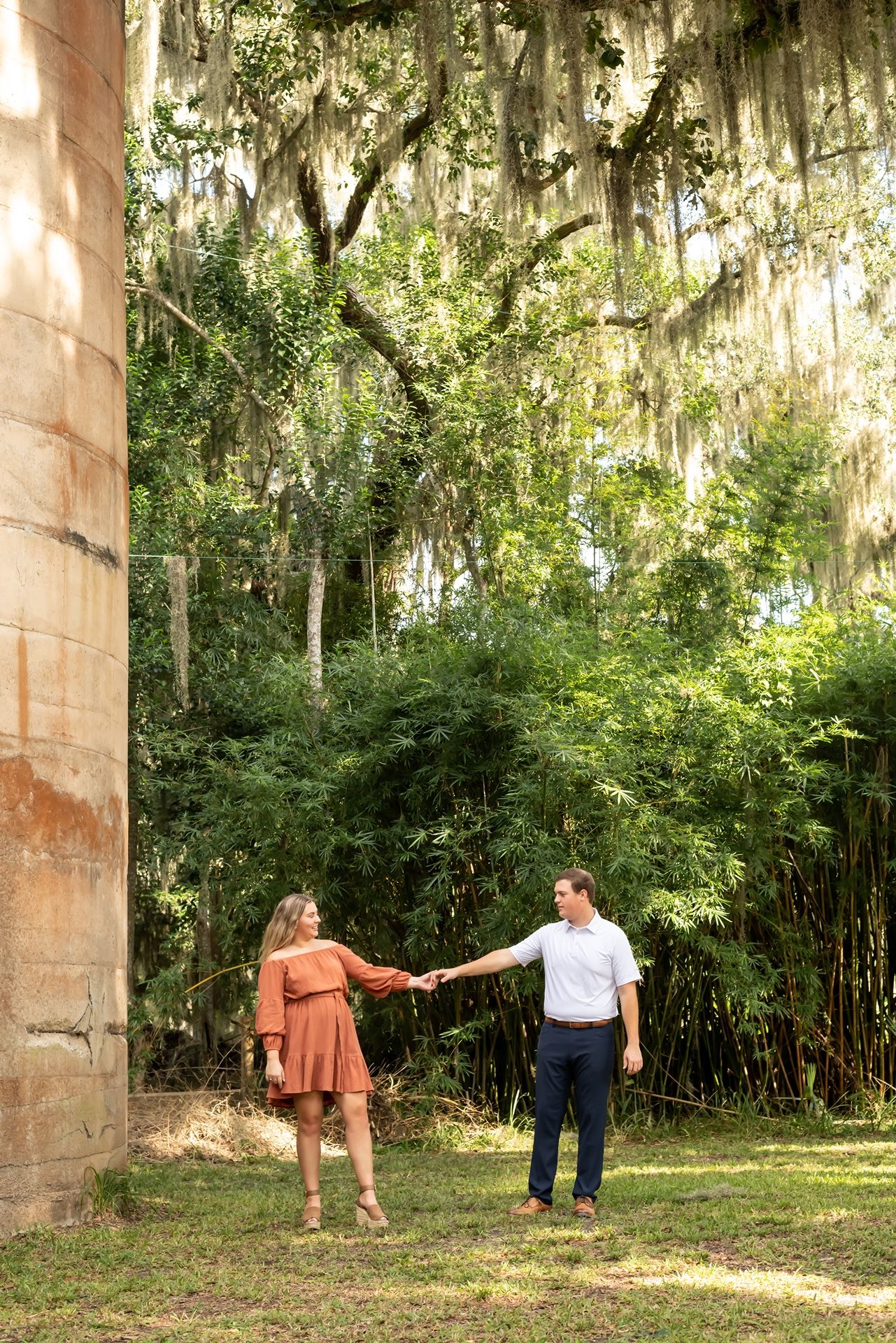 Two people walk together beneath Spanish moss hanging from tall trees in a shaded outdoor setting.