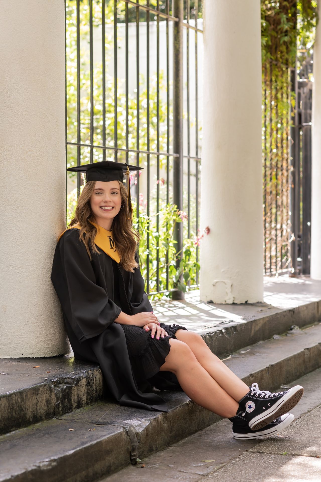 Graduate posing outdoors in black cap and gown with black and white sneakers against a column backdrop with sunlight.