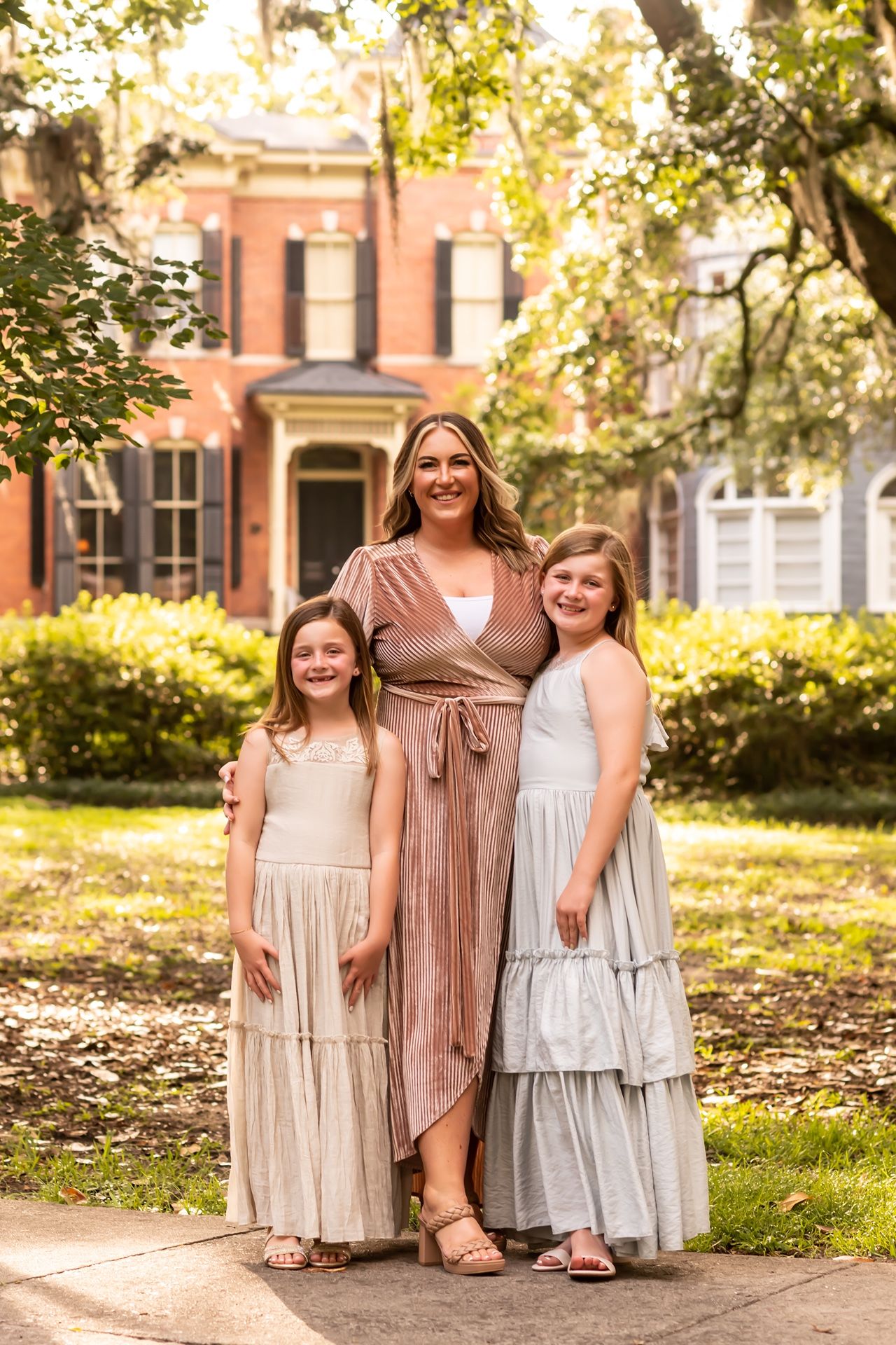 Two young people and an adult pose together in flowing dresses against a historic brick home backdrop.