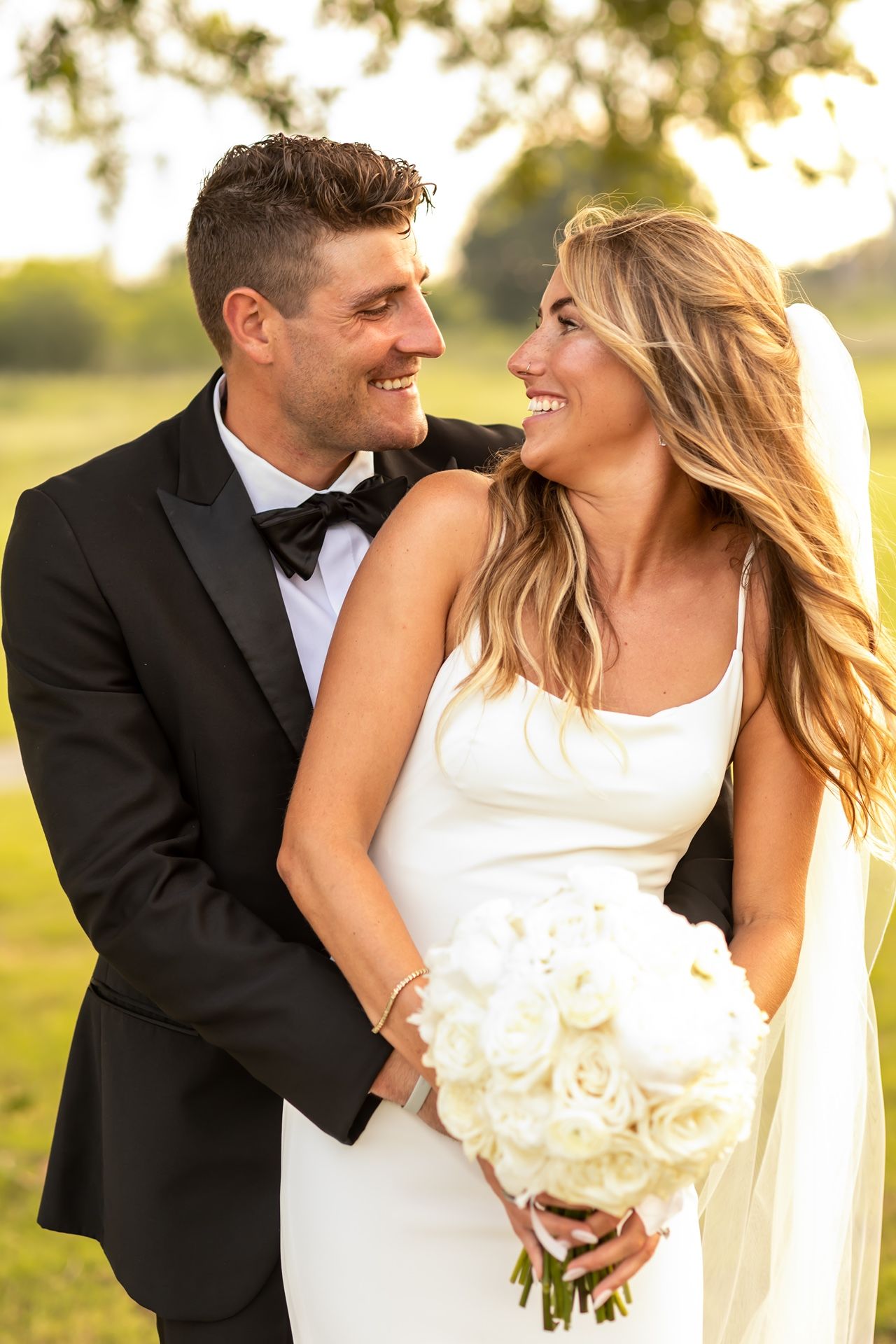 Newlywed couple sharing joyful moment outdoors at sunset with bride holding white rose bouquet.