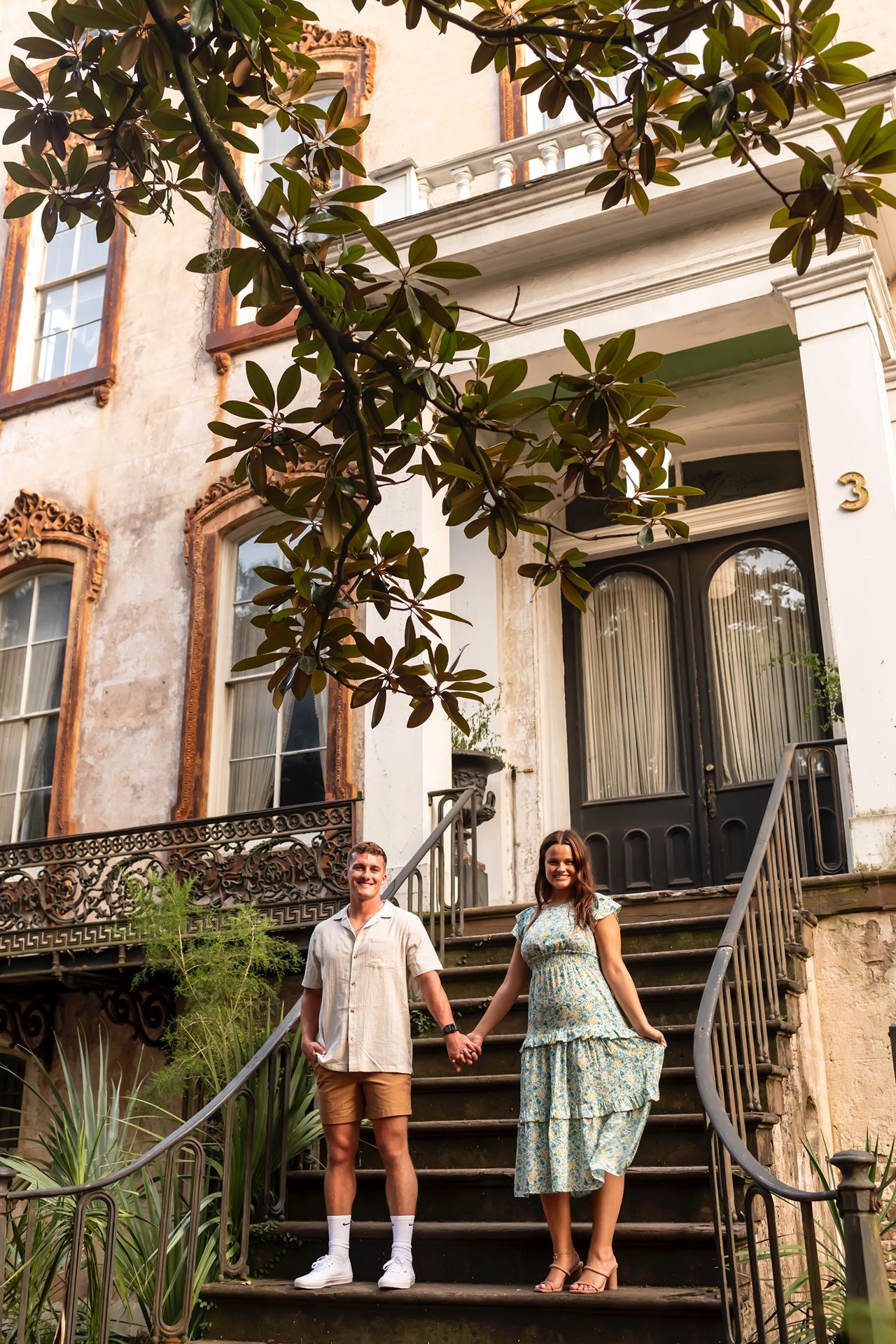 Couple stands together on historic building steps surrounded by lush magnolia branches and greenery.