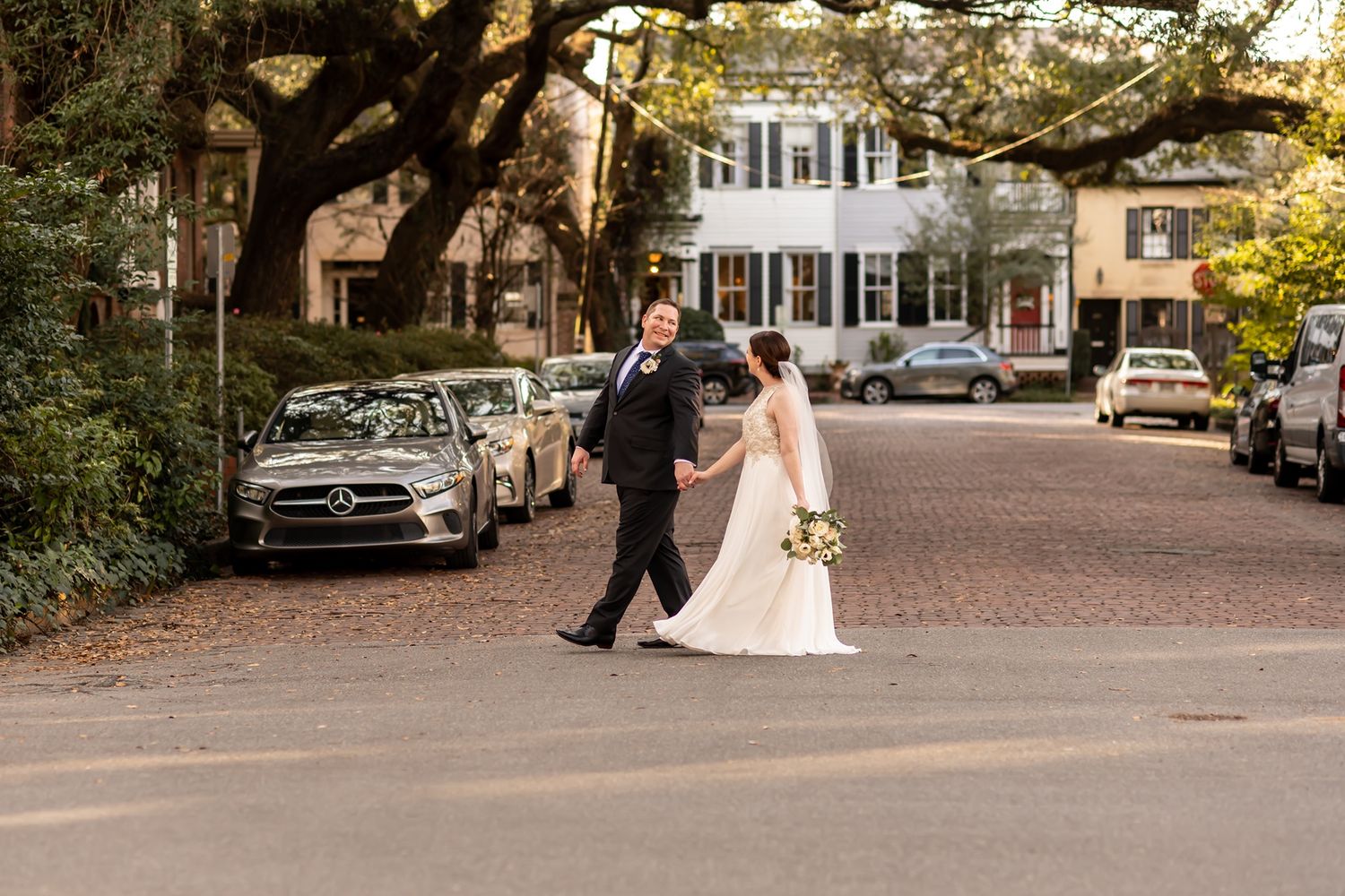 A couple poses during their wedding photo session next to antique cars on a brick-lined street.