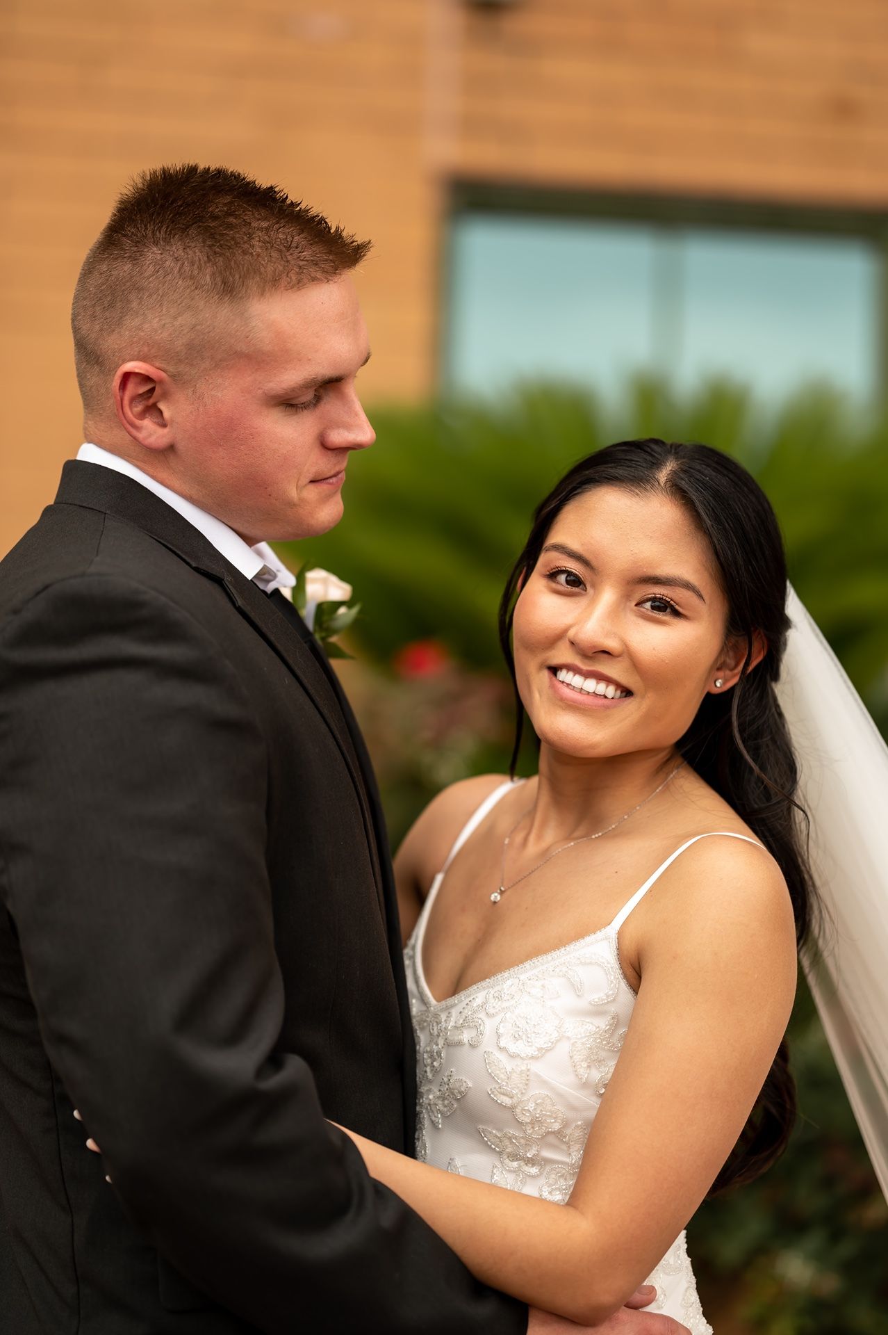 A bride and groom pose together outdoors on their wedding day, with the bride in a white dress and groom in a dark suit.