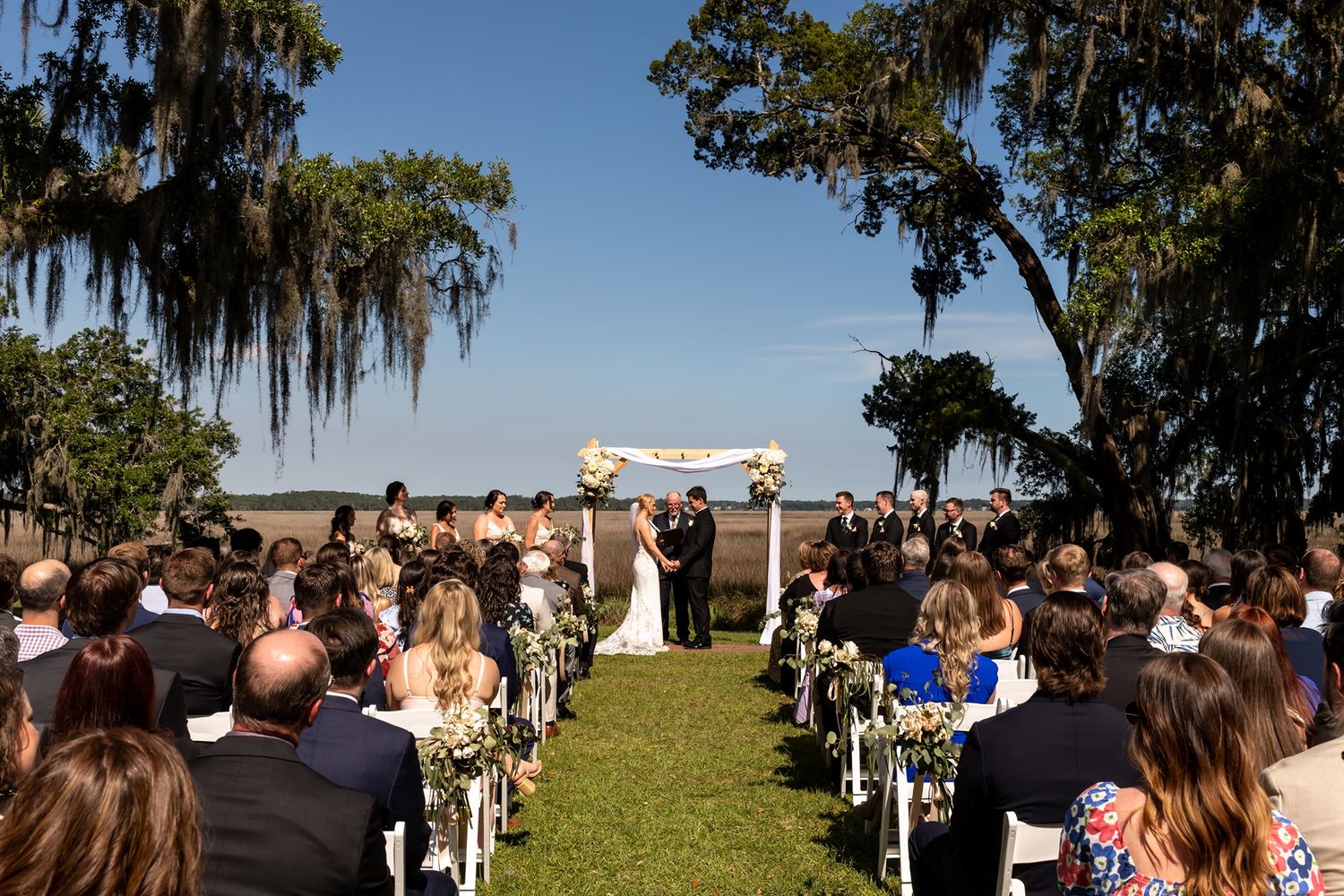 An outdoor wedding ceremony takes place on a grassy lawn with Spanish moss-draped trees and white chairs under blue skies.