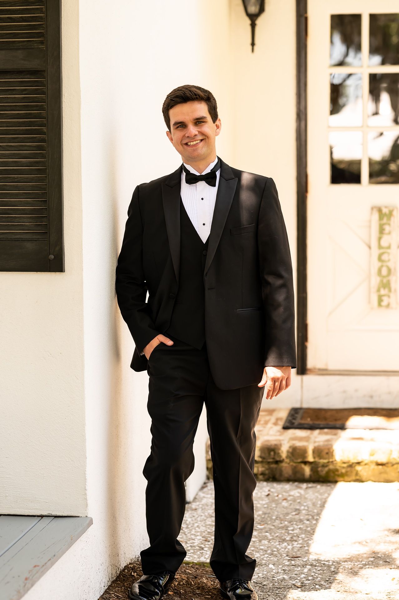 A figure in a classic black tuxedo and bowtie stands confidently against a white architectural backdrop.