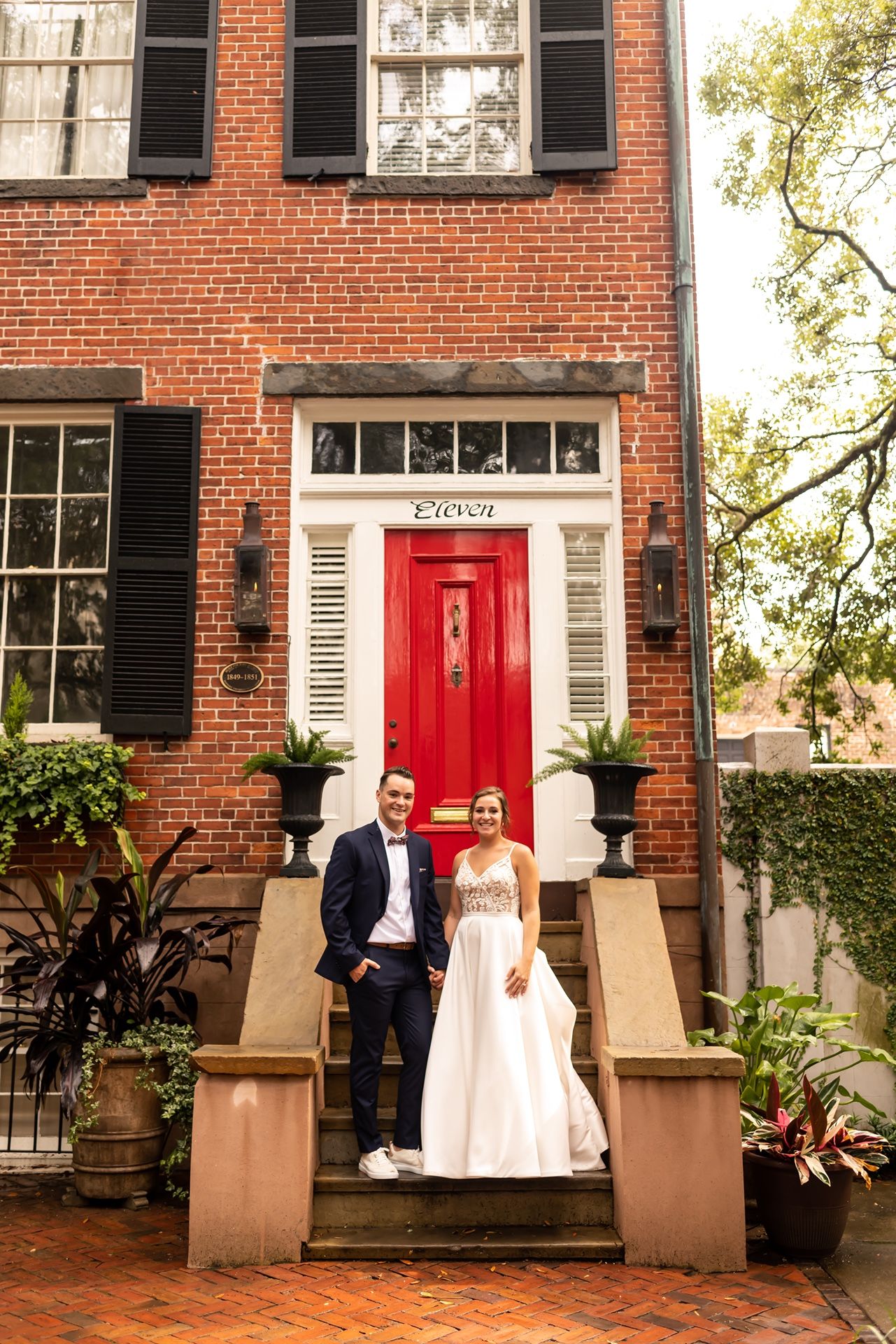 A couple stands on brick steps in front of a historic red brick townhouse with a bright red door and black shutters.