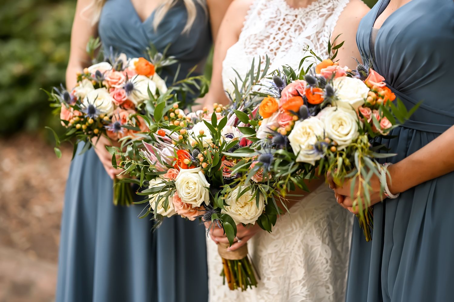 Beautiful wedding bouquets with peach roses, orange blooms and greenery held against dusty blue bridesmaid dresses.
