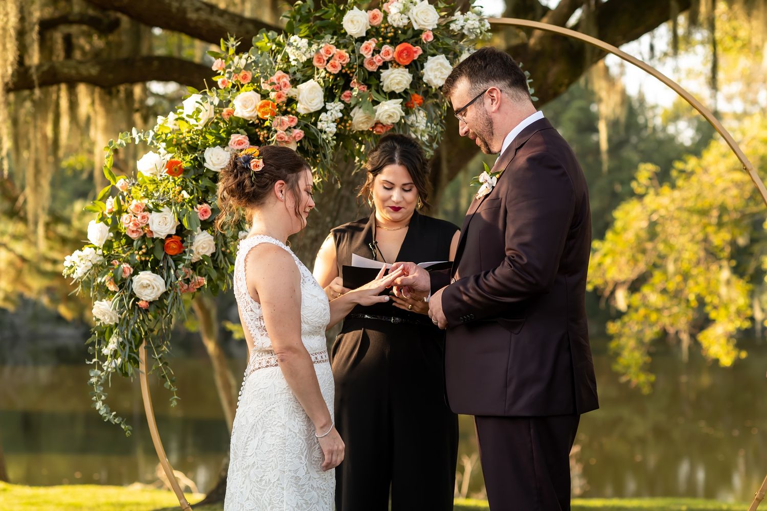 A beautiful outdoor wedding ceremony takes place under a floral arch as the couple exchanges rings at sunset.