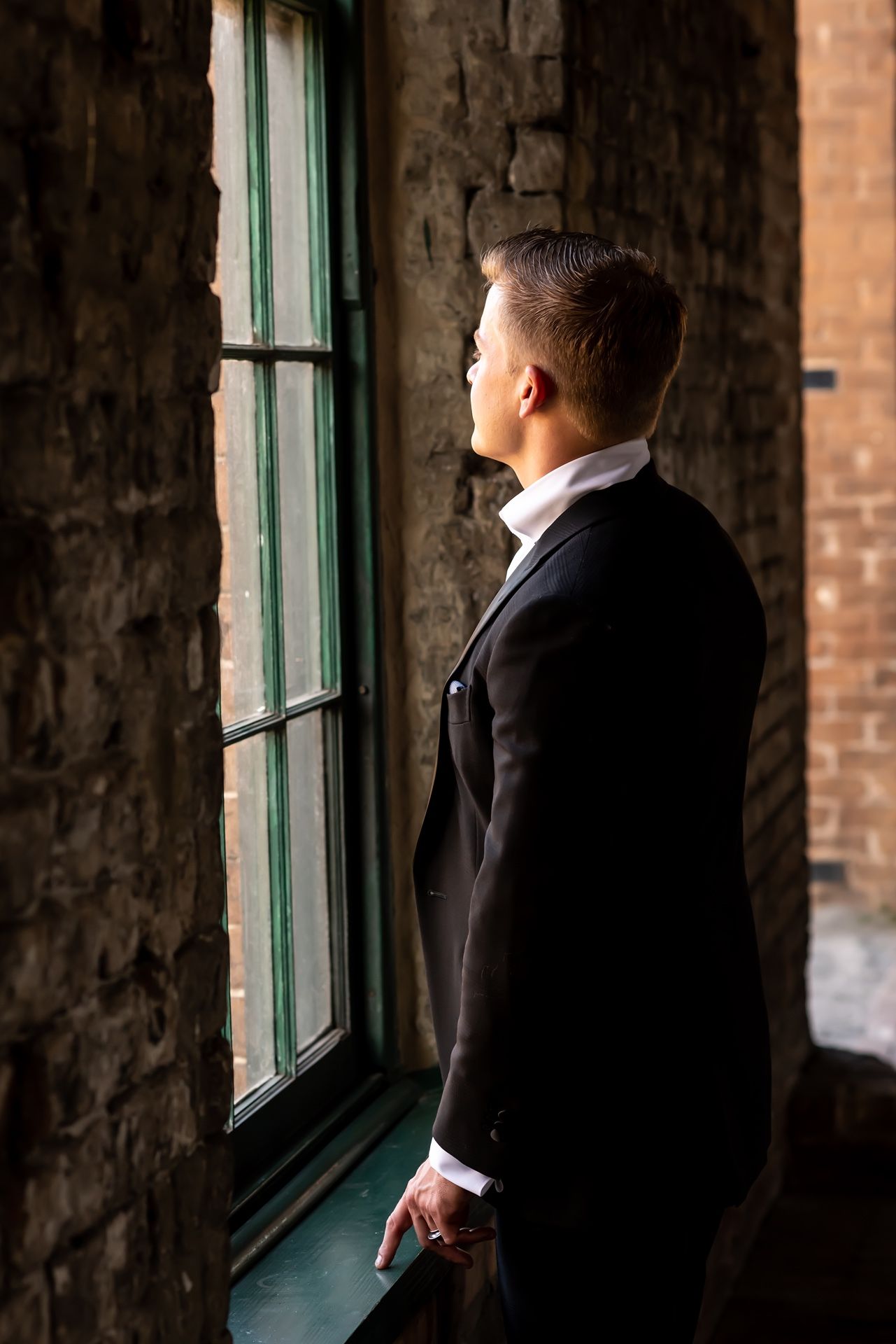 Silhouettes of men in formal attire stand near tall windows in a rustic brick building interior.