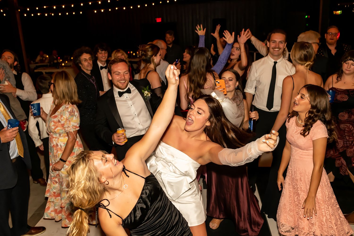 Energetic crowd dances together at a lively party reception with string lights overhead.