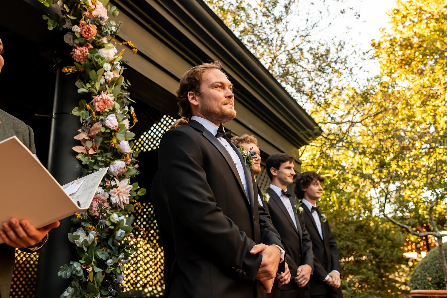 A wedding ceremony scene at sunset with floral decorations and standing figures under a decorative arch.