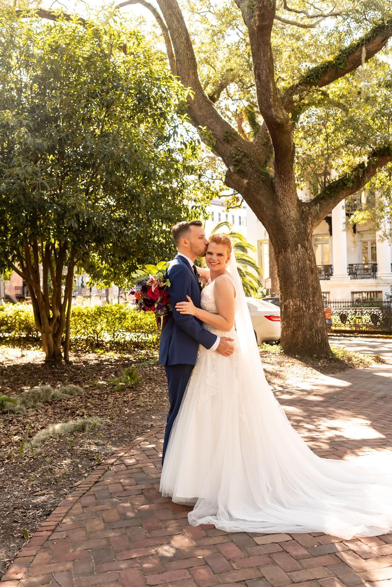 Newlyweds embrace in a romantic garden setting with brick pathways and Spanish moss-draped oak trees in the background.