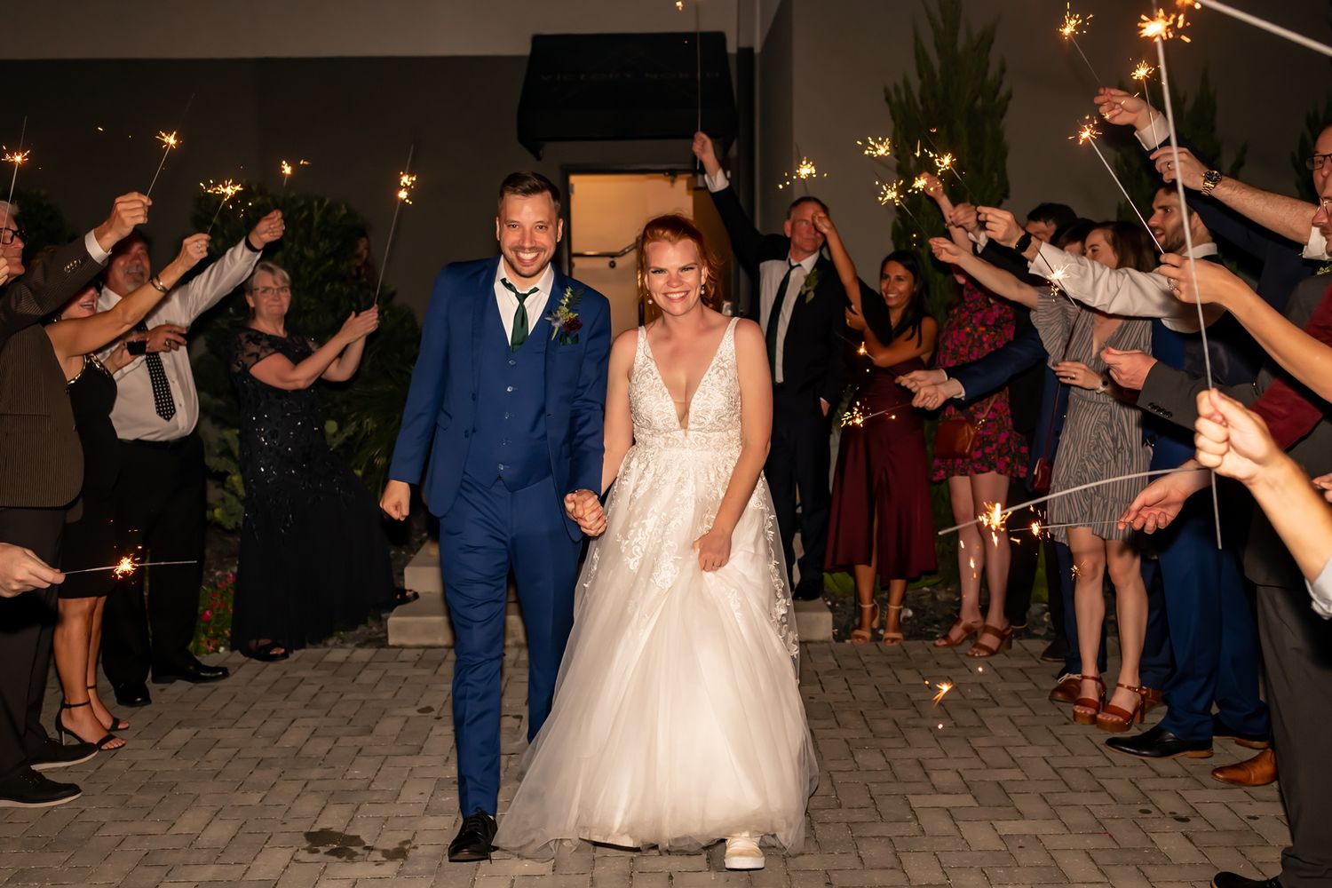 A couple walks through a path lined with their guests holding sparklers at a nighttime wedding celebration.